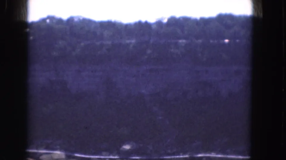 1964 Albany home-movie frame showing a distant tree-covered hillside, pale sky, dark gate edges, and soft blue-purple film color.
