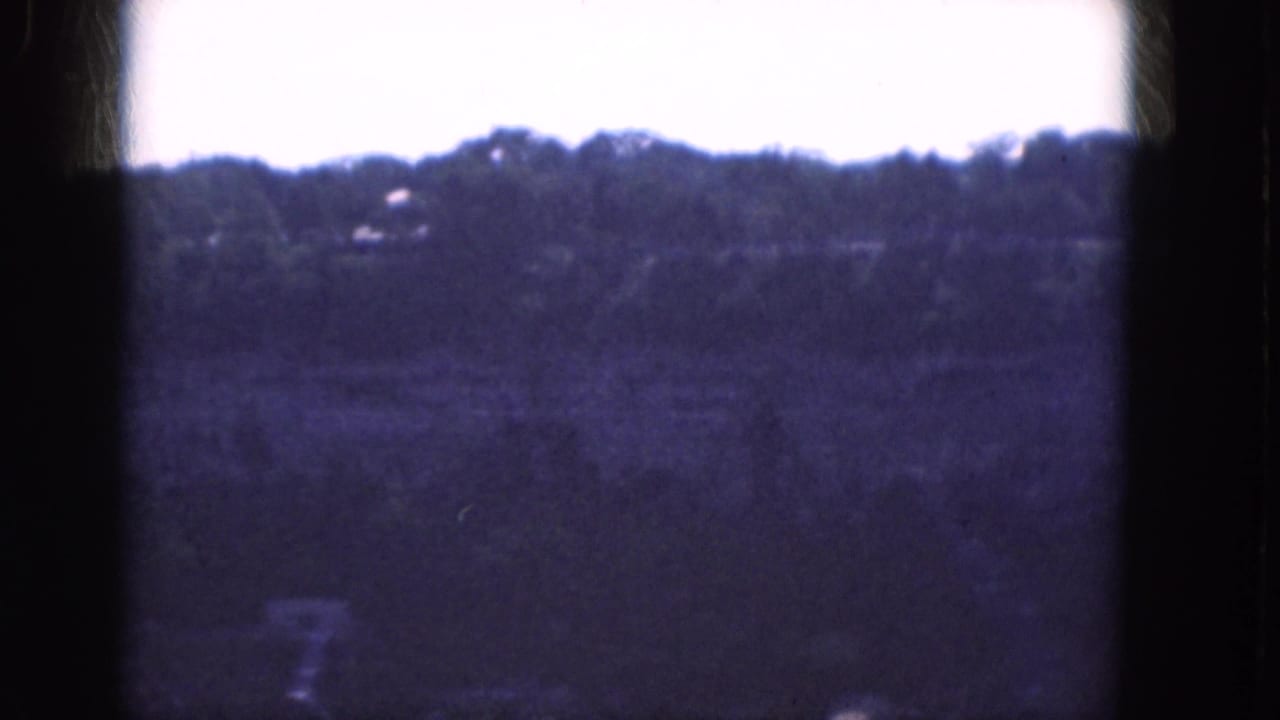 Soft 8mm view of a distant wooded ridge with faint structures near the treeline.