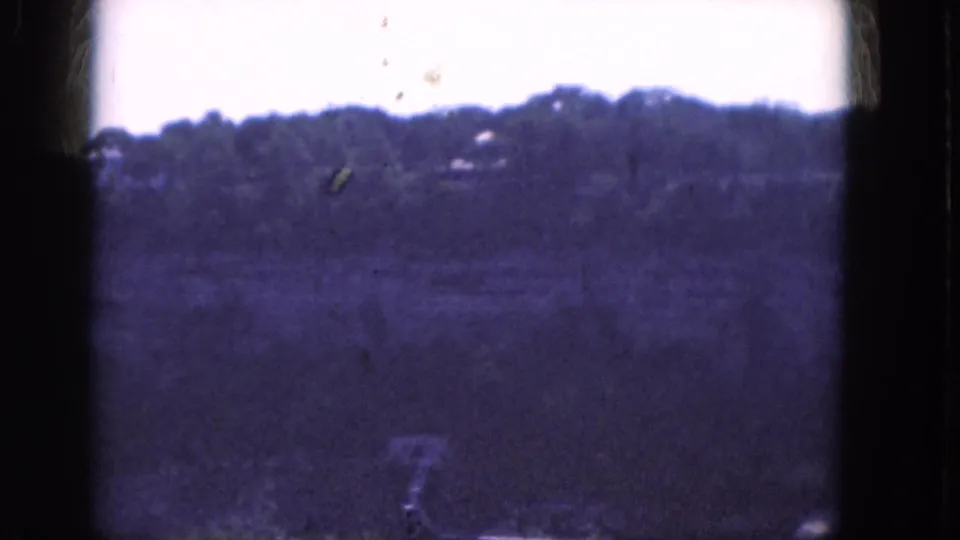 1964 Albany home-movie frame showing a wooded hillside, pale sky, open foreground, dust specks, and a small pale object near the bottom edge.