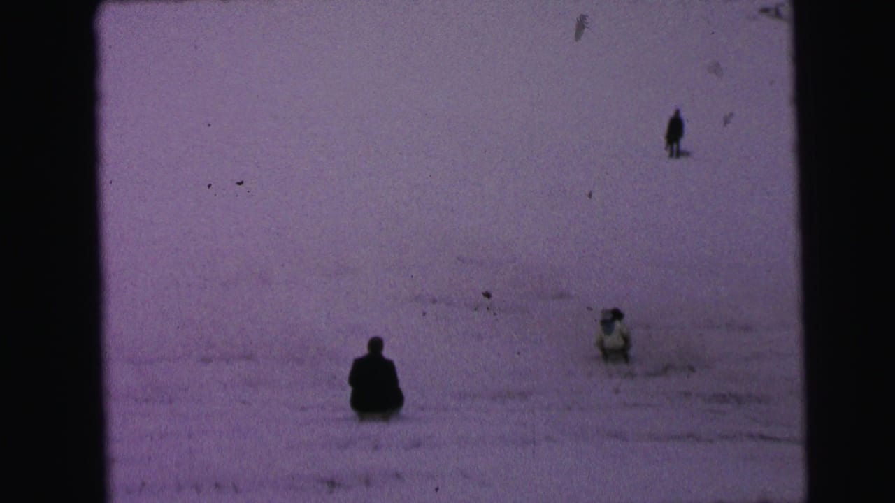 Several people sled or stand on a wide snowy hill with dark film borders.