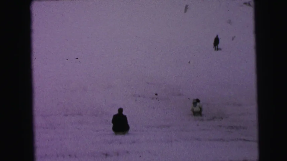 1964 Camden home-movie frame showing distant figures on a snow-covered slope, including sledders and one standing person.
