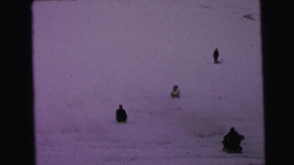 1964 Camden footage frame with several small figures sledding or sitting on a broad winter hillside.