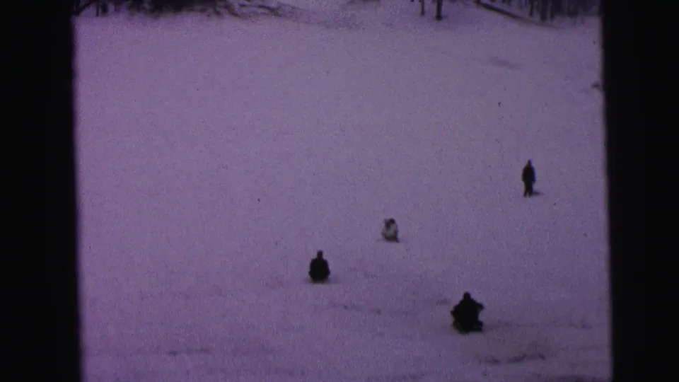 1964 Camden winter home-movie frame showing people sledding on a snow slope with trees faintly visible near the top edge.