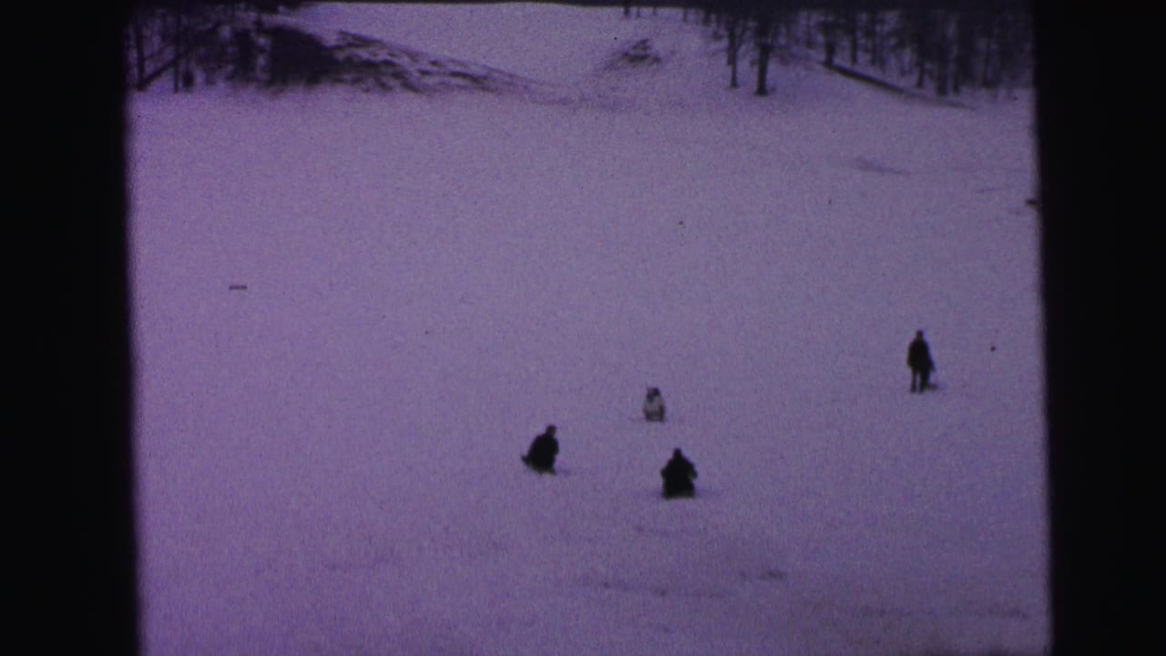 1964 Camden home-movie frame showing distant figures on a snowy hillside, with bare winter trees and dark 8mm film borders.