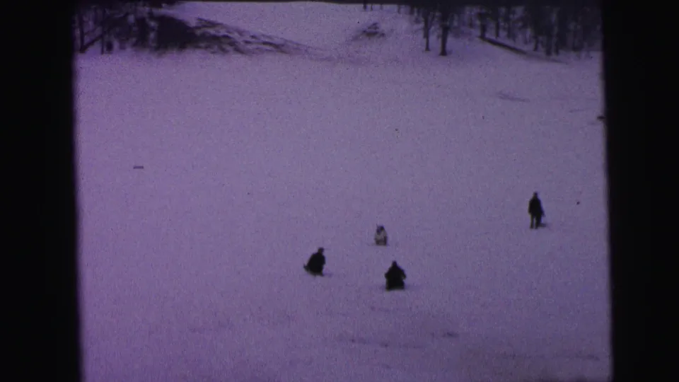1964 Camden home-movie frame showing distant figures on a snowy hillside, with bare winter trees and dark 8mm film borders.