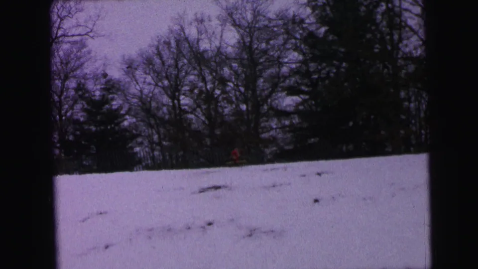 1964 Camden home-movie frame of a snowy hillside below leafless trees, with a small red figure partly visible near the background.