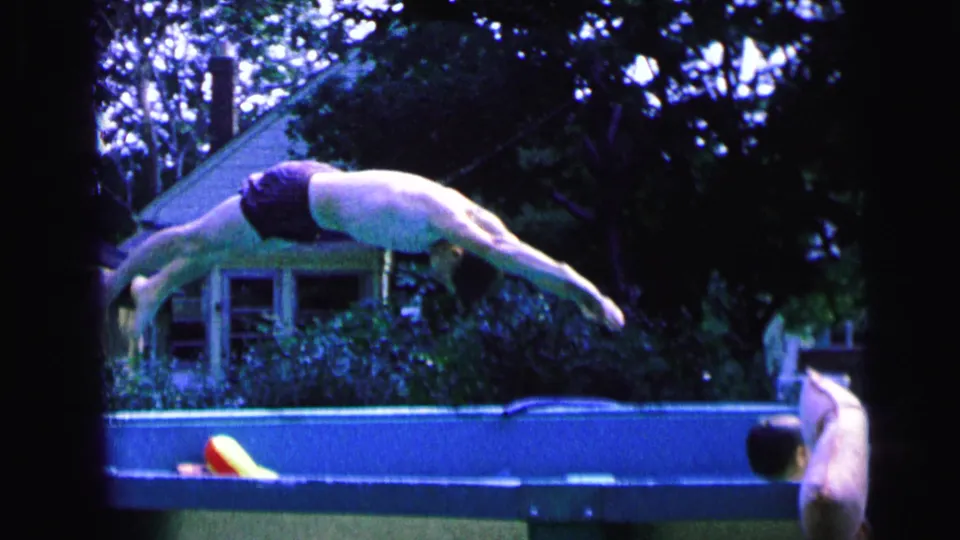 The man is airborne in a straight dive over the backyard pool, with a house and trees in the background.