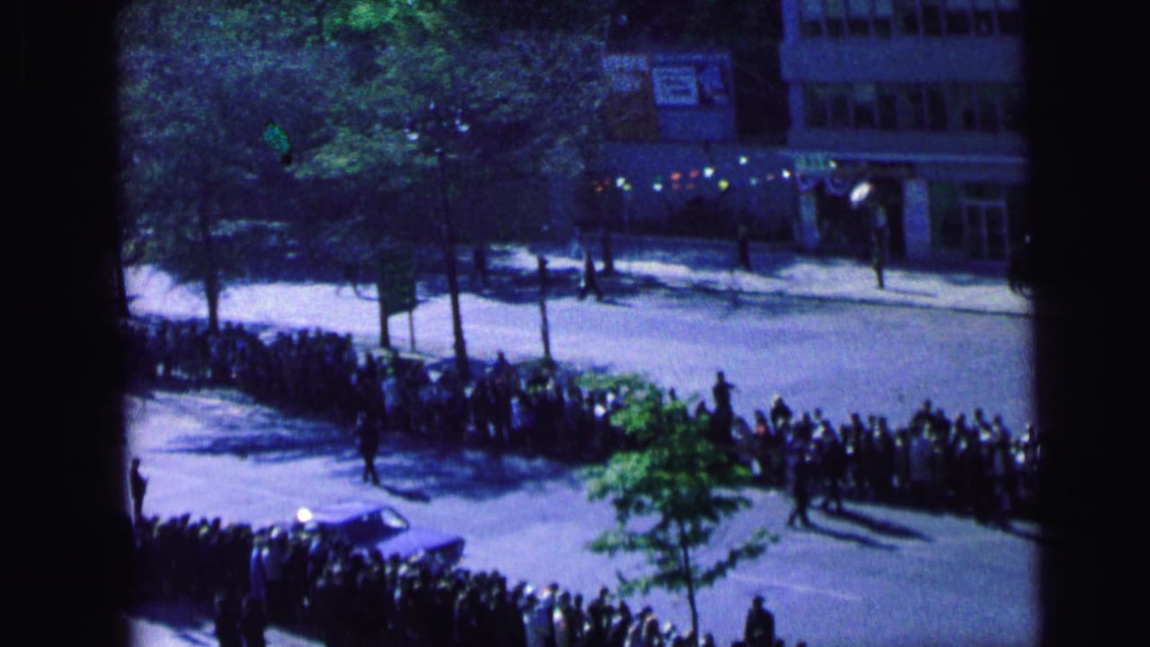 Elevated 1964 Camden home-movie view of people gathered along sidewalks beside trees, buildings, and a parked car.