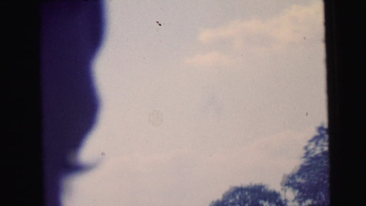 1964 Camden 8mm frame with a dark elongated shape beside a large blue blur against pale sky, with faint clouds and tree tops at the lower edge.