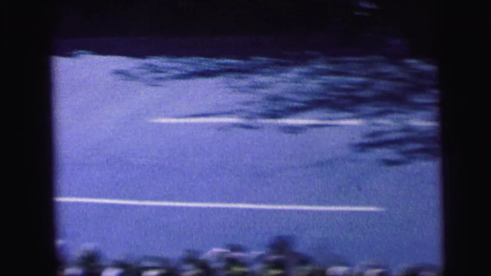 A quiet portion of the Camden street appears between moving vehicles, showing lane markings, shadowed pavement, and handheld 8mm framing.