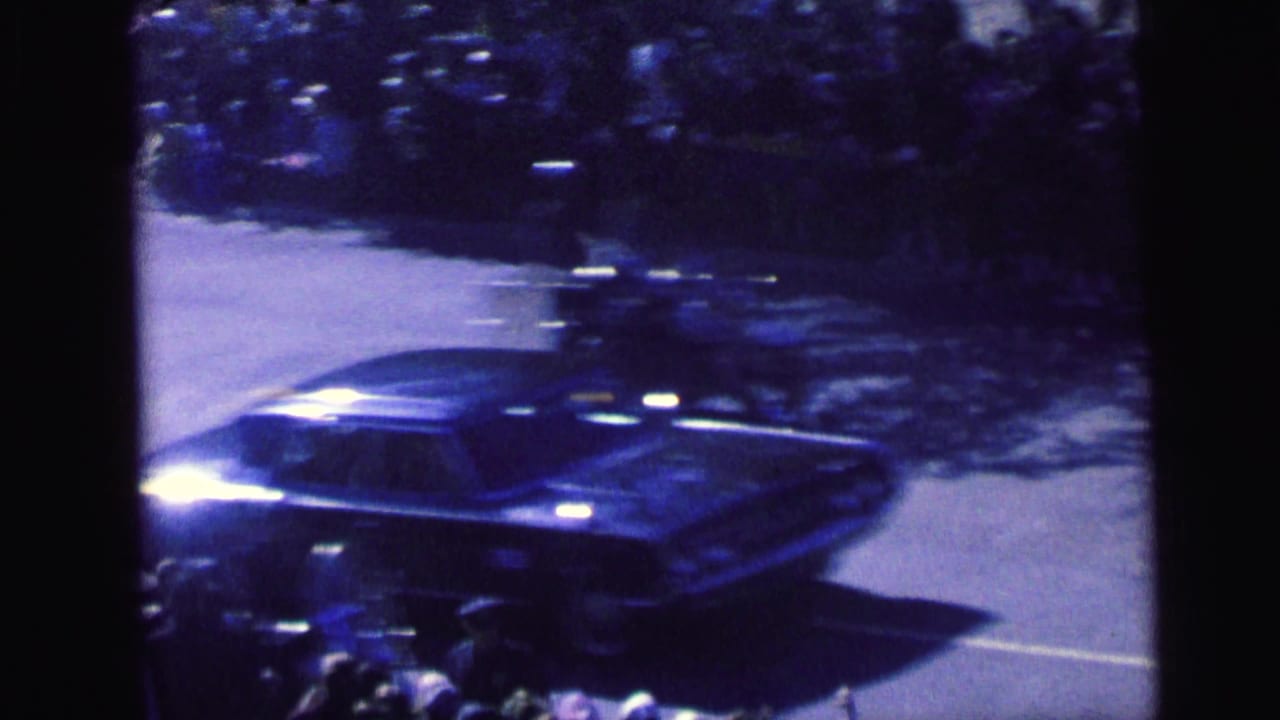 A dark 1960s sedan passes a crowd-lined Camden street, with spectators and possible bicycles or motorcycles visible near the curb.