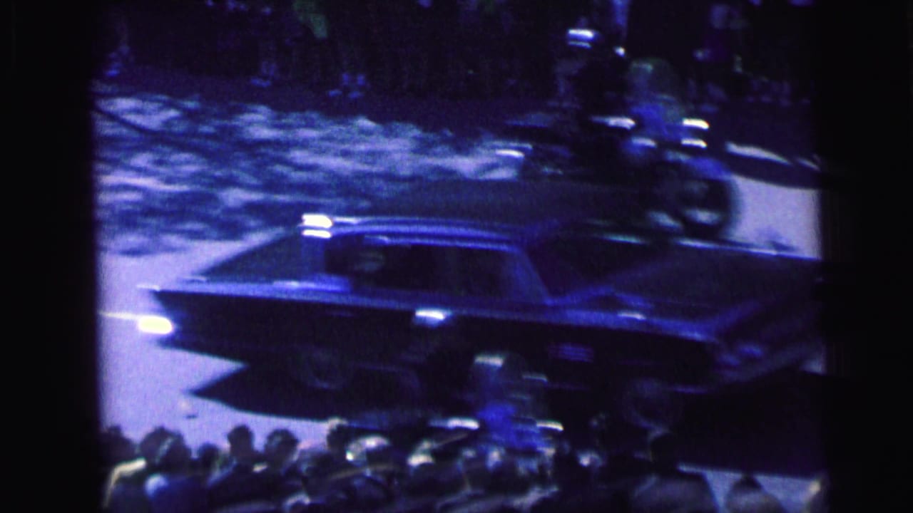 A dark sedan moves past roadside spectators in Camden while a helicopter appears overhead, captured in grainy blue-tinted 8mm footage.