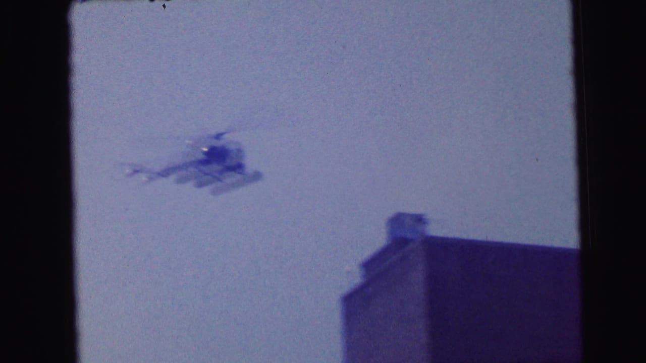 1964 Camden 8mm frame showing a blurred helicopter moving across a pale blue sky above a dark urban rooftop edge.