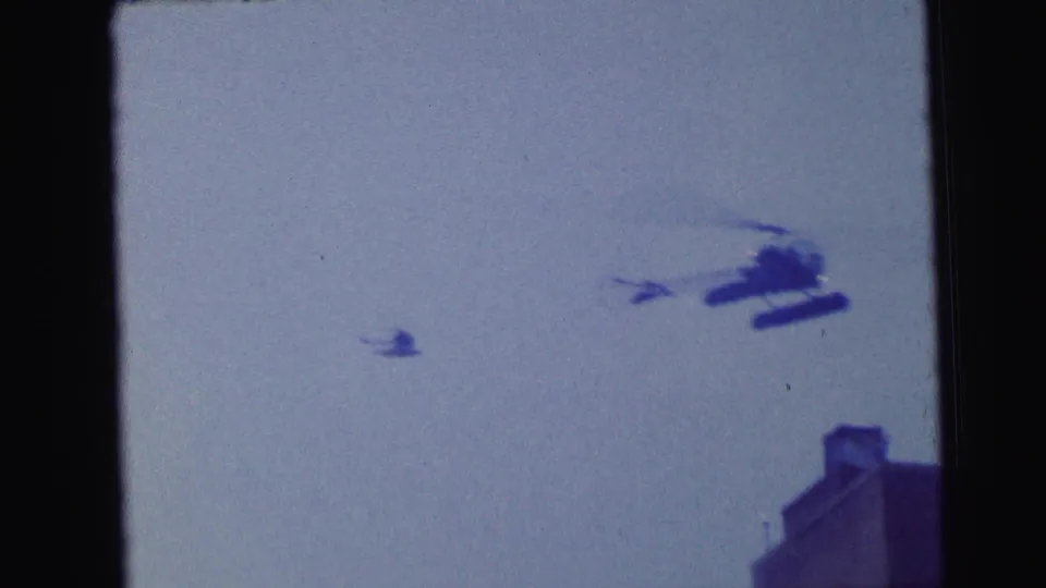 1964 Camden 8mm frame with a helicopter near the upper right and a smaller blurred aircraft shape against the sky over a rooftop corner.