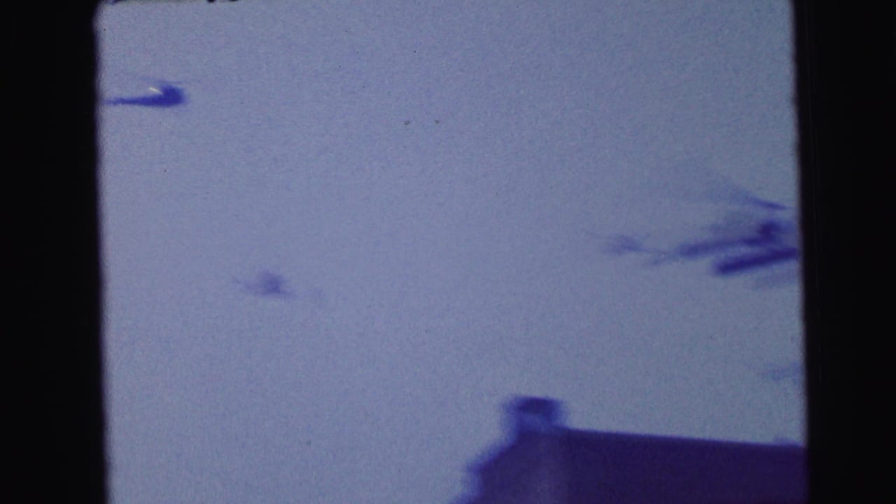 Blue-tinted 8mm frame from Camden showing blurred helicopter motion across the sky, with a dark roof edge at the lower right.
