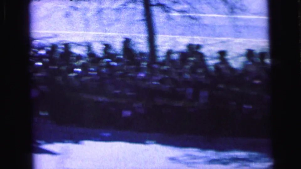 Distant spectators line a sunlit Camden street in a heavily blurred 1964 home-movie frame shot through a screen or window.