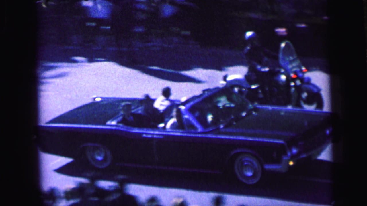 A dark convertible in a Camden motorcade passes below the camera, with motorcycle escorts and spectators along the street.
