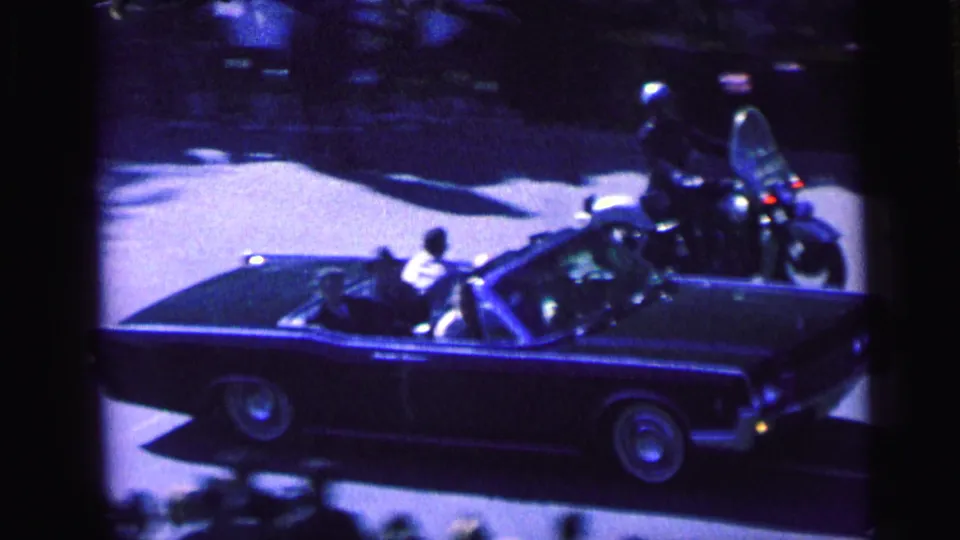 A dark convertible in a Camden motorcade passes below the camera, with motorcycle escorts and spectators along the street.