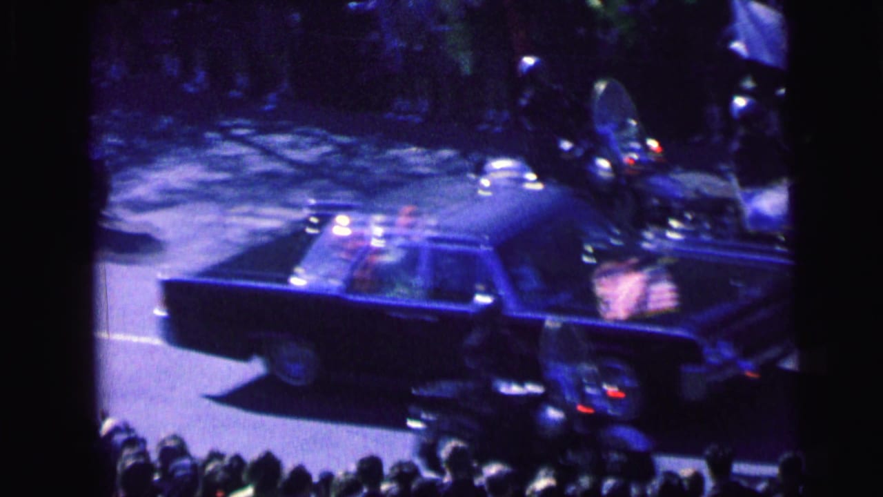 The motorcade continues through Camden as the dark convertible and motorcycle escorts pass beneath an elevated, screened viewpoint.