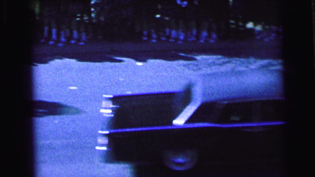 Dark convertible moving past spectators along a Camden street during the 1964 motorcade, seen in grainy blue 8mm footage.