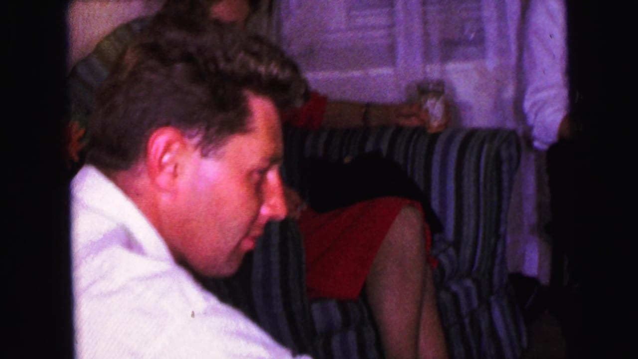 Man in a white shirt sits in profile beside a striped couch in a dim living room.