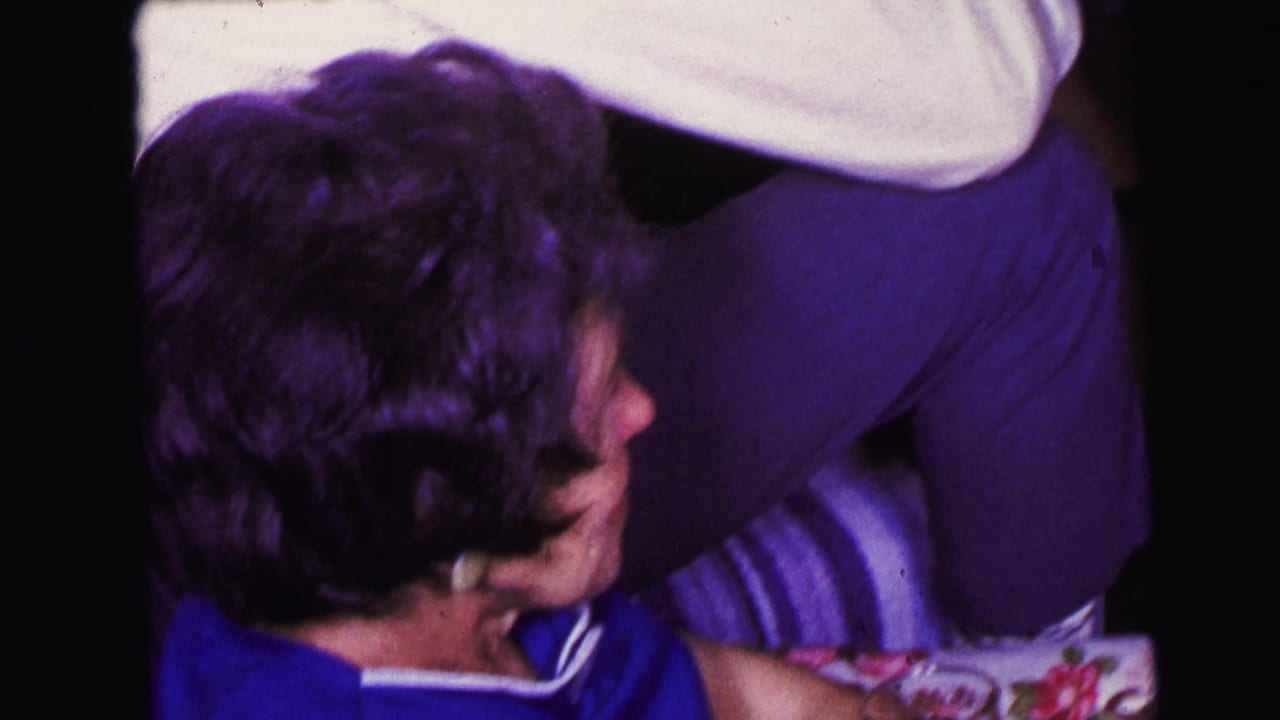 Older woman in a blue top sits near another adult standing in purple pants.