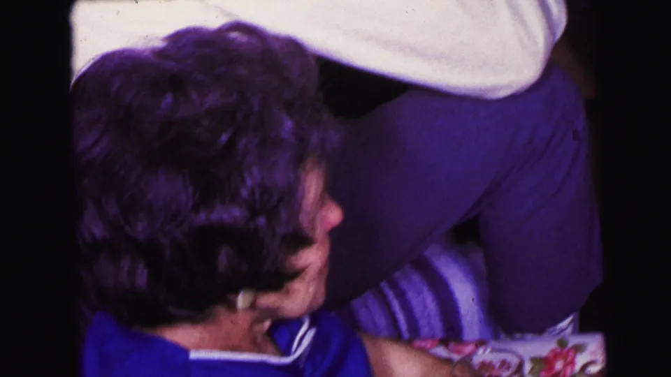 1964 Camden home-movie frame with an older woman in a blue top seated on a couch while another adult stands close by in a private room.