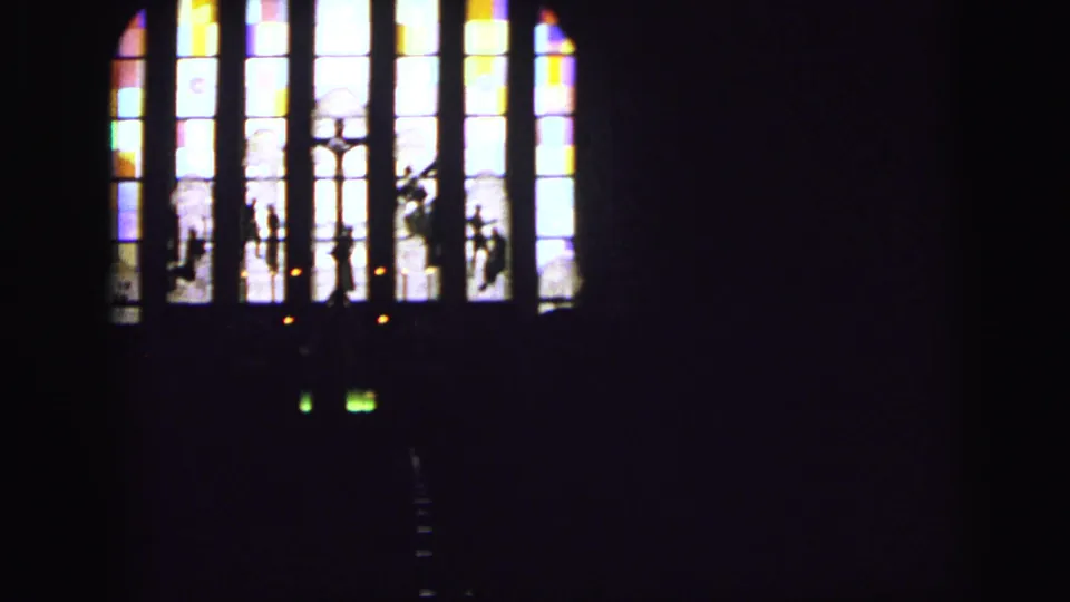 Dim 1964 Camden church interior with stained-glass panels, possible religious figures, and cross forms visible in the window.