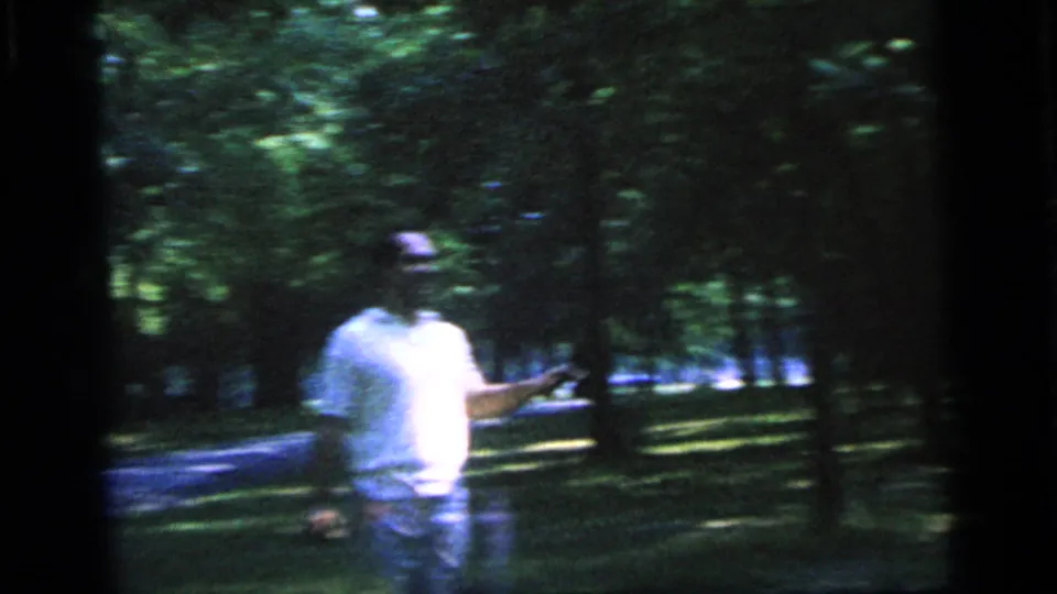 A man in a white shirt extends one arm during the 1964 Camden park badminton scene, with trees and shaded grass behind him.