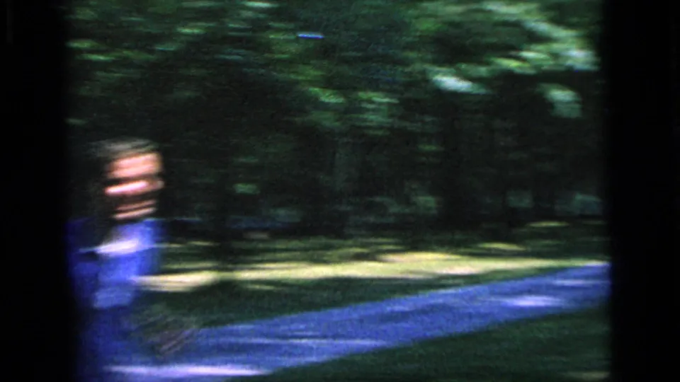 A blurred man in a blue shirt appears near a curving park path in the 1964 Camden badminton footage, with dense trees behind him.