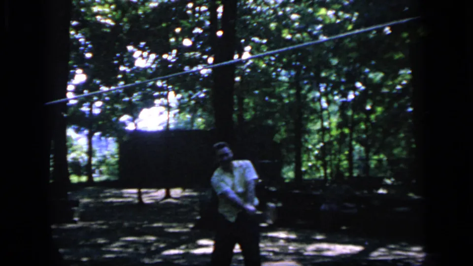 A man stands in a shaded wooded clearing near a badminton net, with strong contrast and 8mm grain in the 1964 Camden footage.
