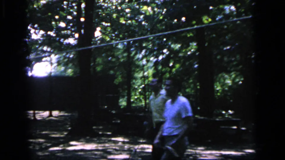 A 1964 8mm frame shows adults in casual clothing moving through a Camden wooded clearing while playing badminton.