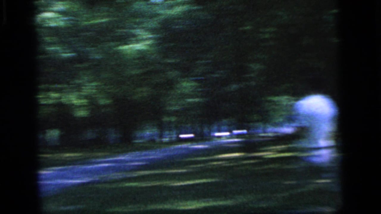1964 Camden home-movie frame showing a tennis player in white moving through a wooded park setting.