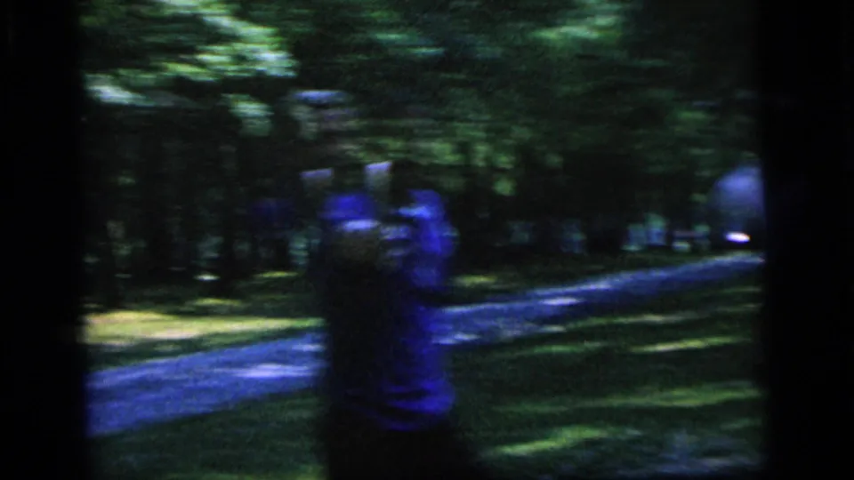 1964 Camden frame showing a player in a blue shirt near a wooded park path during casual tennis play.
