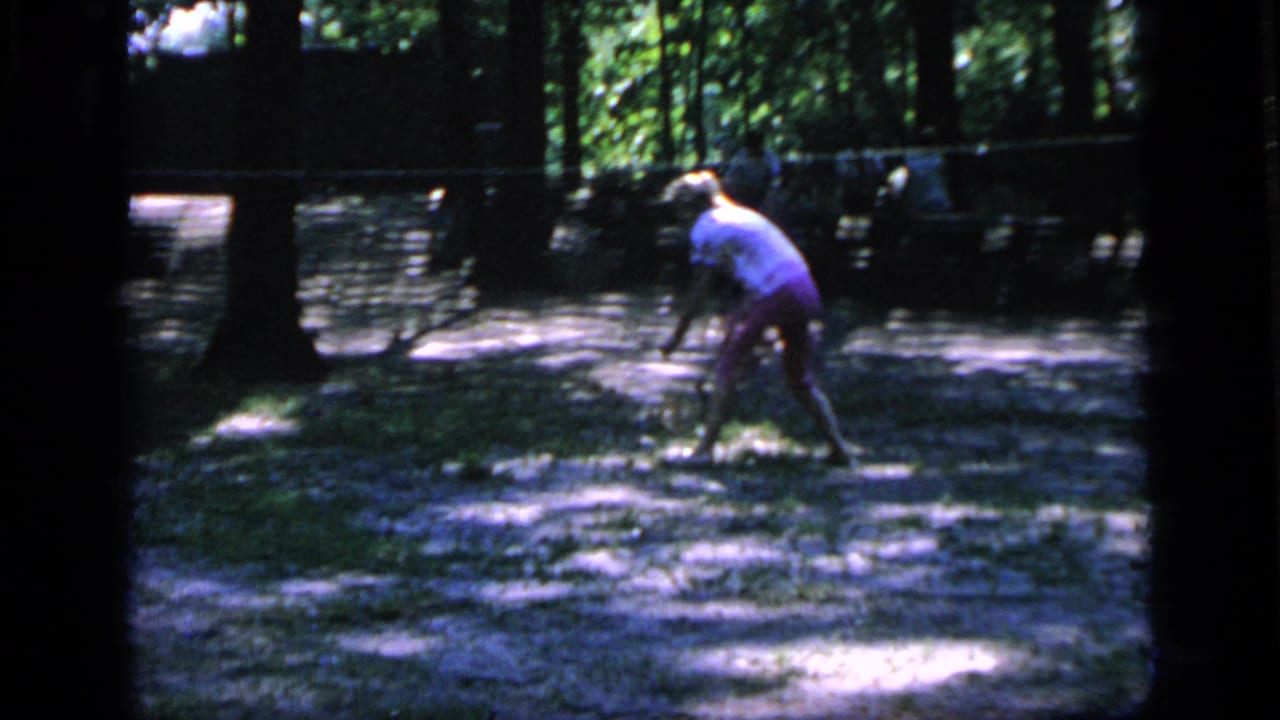 In 1964 Camden, New Jersey footage, a woman in a light top bends with a badminton racket under trees.