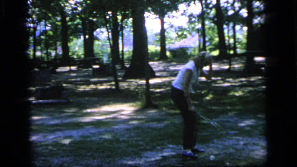 A woman in a white top stands ready with a badminton racket in a wooded Camden park scene.