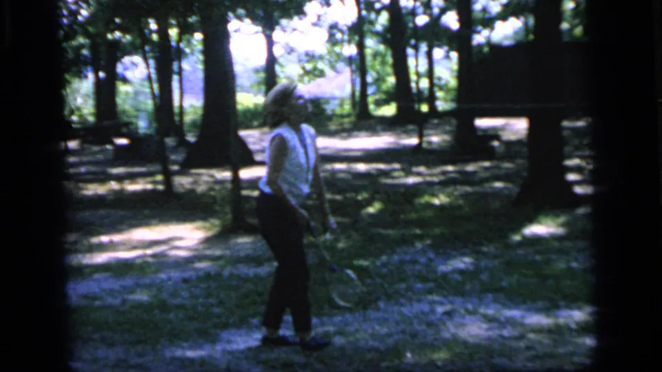 The 1964 Camden home-movie frame shows the woman holding a racket in a wooded recreation area.