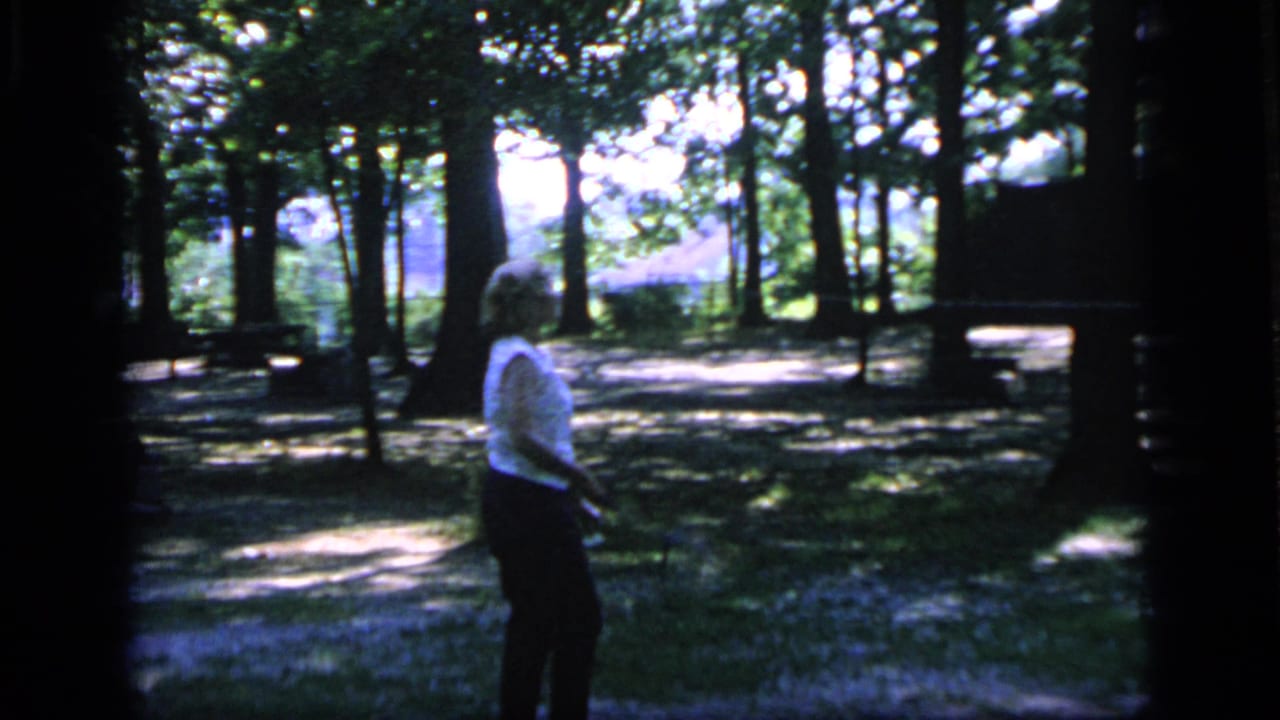 In 1964 Camden home-movie footage, a woman stands among trees in dappled light with a badminton racket near a simple outdoor play area.