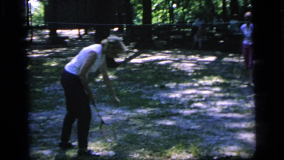 A woman bends to reach or retrieve the shuttle near a makeshift badminton net, with another player visible in the background.