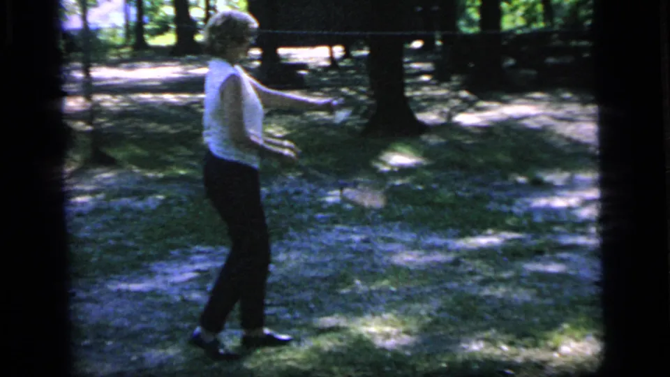 The woman is seen in profile with her racket extended in the 1964 Camden garden badminton scene, surrounded by trees and mottled sunlight.