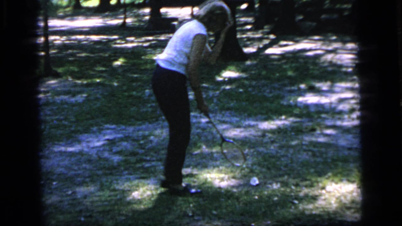 Woman bends with a badminton racket near a shuttlecock on the ground.