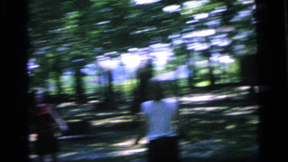 A motion-blurred 8mm frame shows people moving during badminton play in a shaded Camden park in 1964.