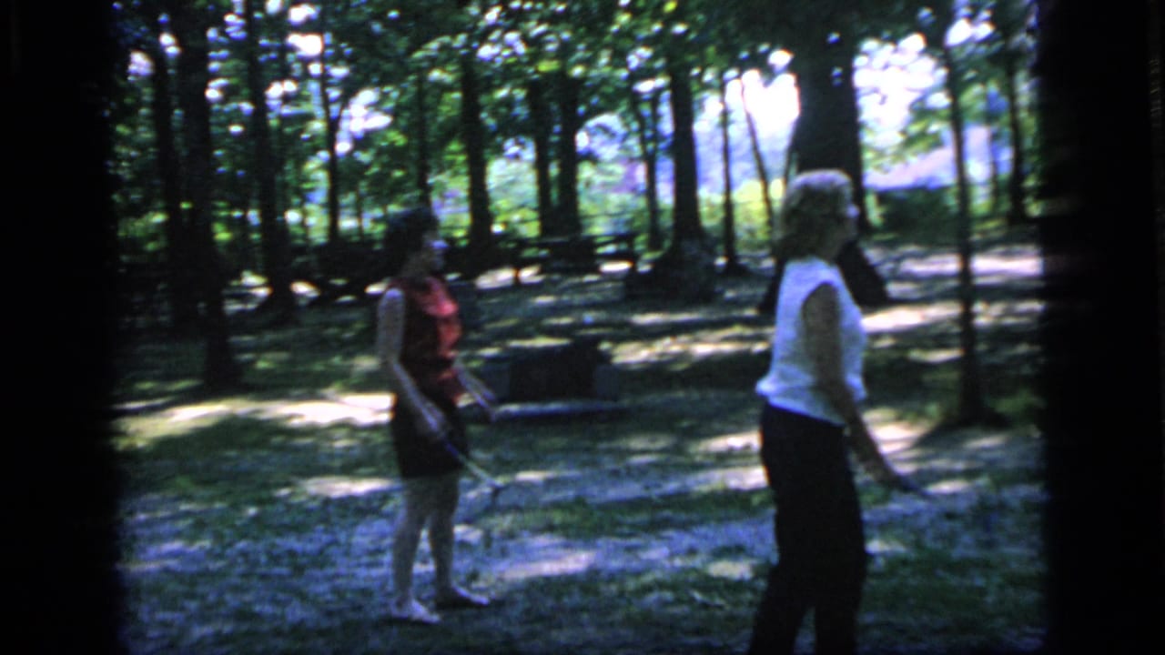 Woman in a red sleeveless top stands with a racket while another woman walks nearby in the park.