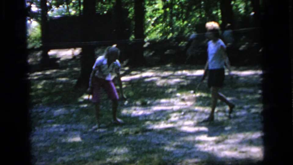 Two women with rackets pause or reset during a casual 1964 badminton game in a shaded Camden park clearing.