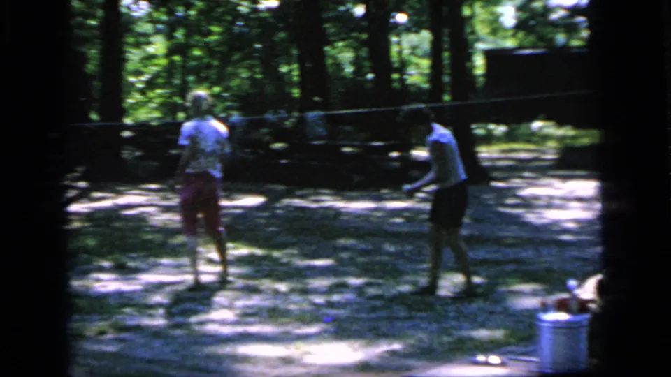 Two women face one another with badminton rackets in a Camden park, with a pail and shaded trees visible nearby.