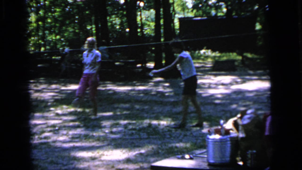 In 1964 Camden, New Jersey home-movie footage, two women play badminton in a wooded park with picnic items nearby.