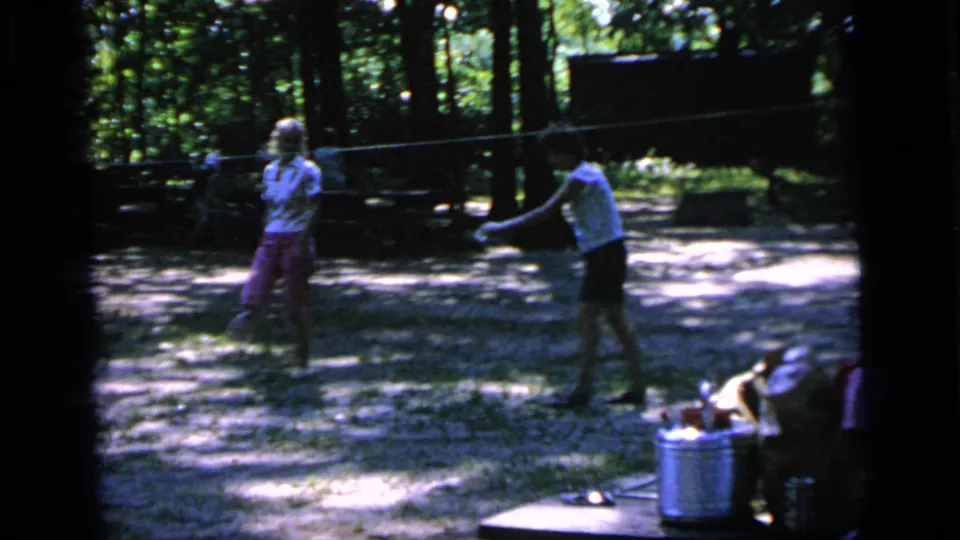 In 1964 Camden, New Jersey home-movie footage, two women play badminton in a wooded park with picnic items nearby.