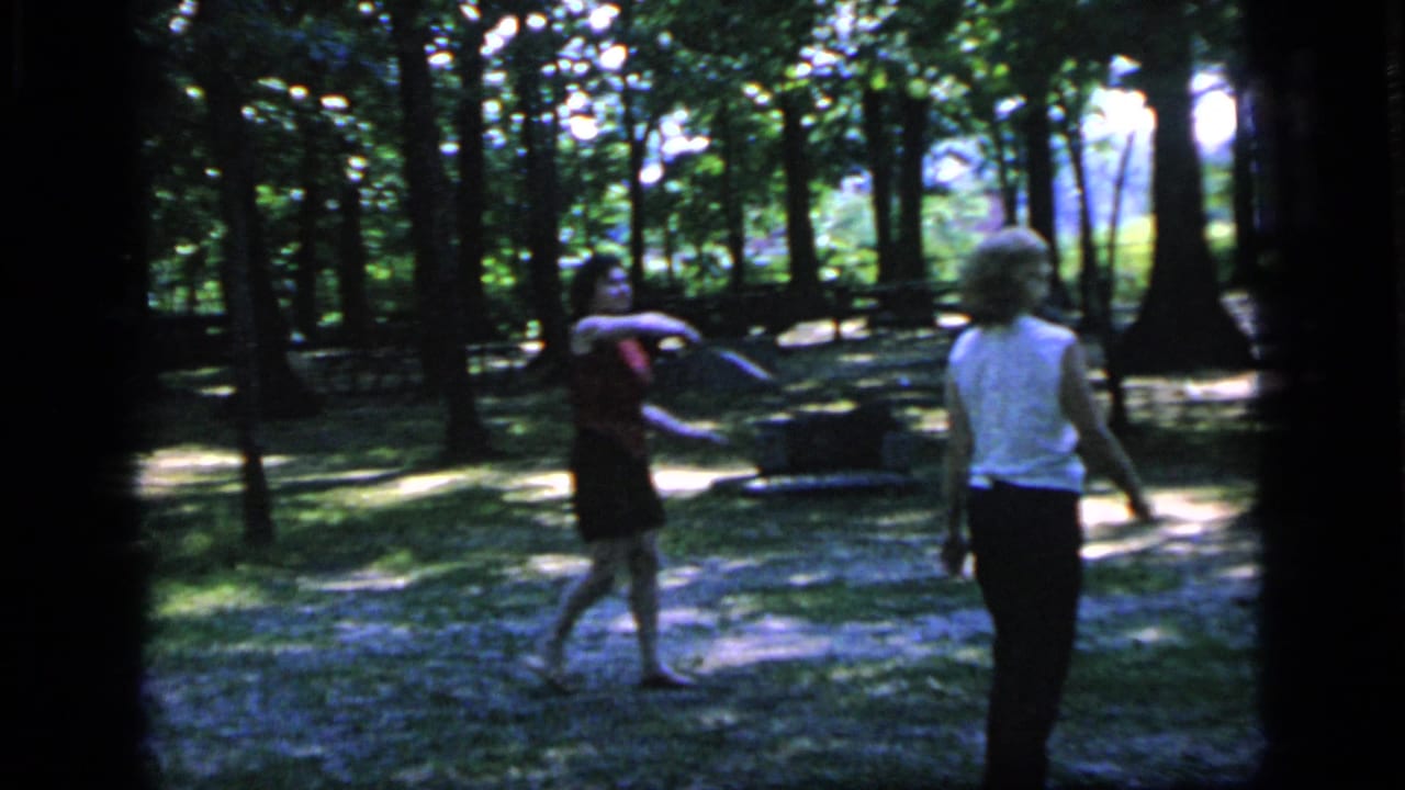 Two women move across a wooded park while playing badminton.