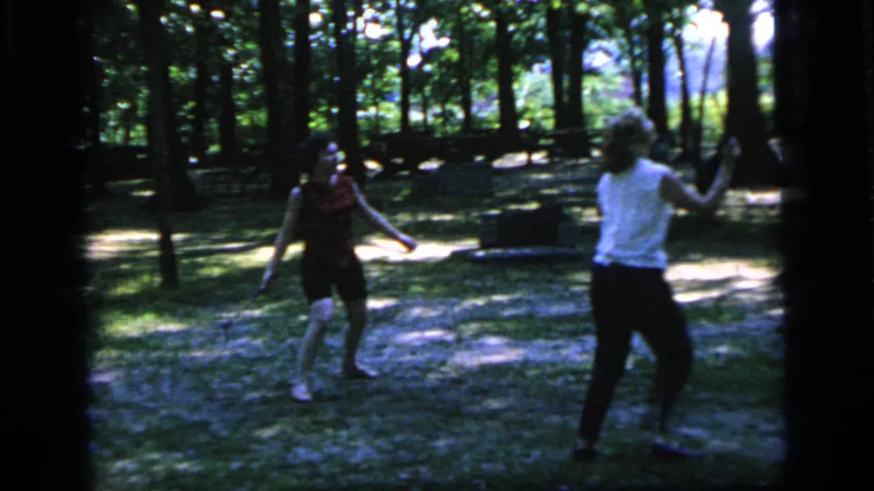 A woman in a red top and a woman in a white sleeveless shirt face each other during badminton in a wooded Camden park.