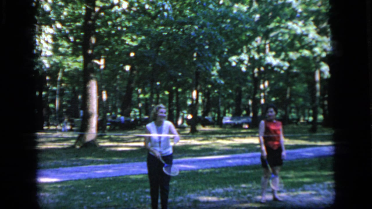 Two women face the camera with badminton rackets in a tree-filled park.